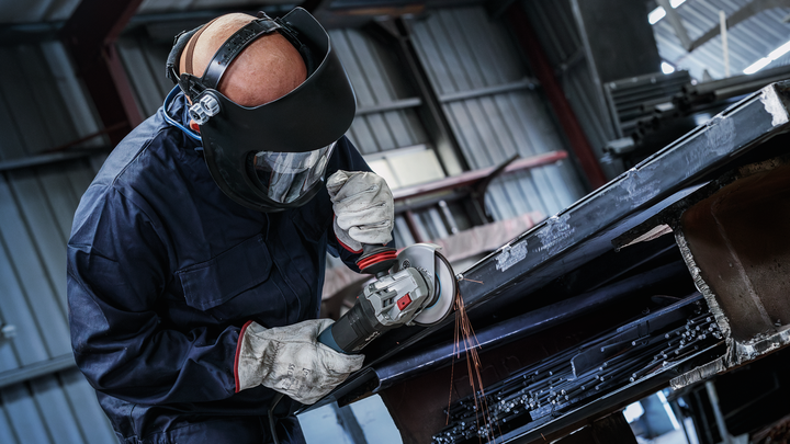 A worker wearing safety equipment grinds metal, creating sparks in an industrial setting.