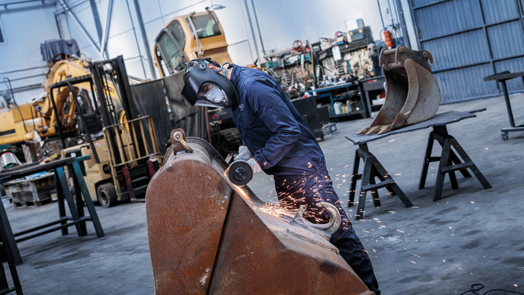 A person wearing safety equipment grinds metal, producing sparks in an industrial workshop.