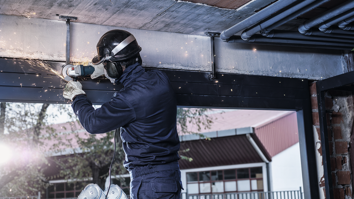 Person wearing safety equipment grinds a metal beam, creating sparks indoors.