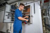 Person wearing safety equipment punches a hole in an electrical cabinet using a cordless tool.