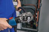 A person uses a cordless hydraulic crimping tool on a thick cable near electrical panels.