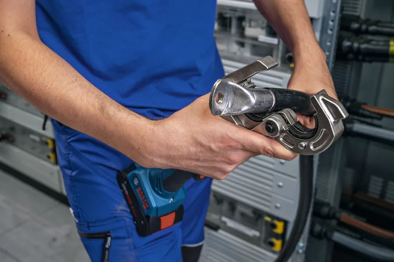 A person wearing safety equipment uses a cordless hydraulic crimping tool on a cable.