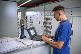 Person operates a laptop connected to a cordless hydraulic crimping tool in an electrical control room.