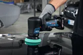 A person wearing safety equipment uses a cordless polisher on a car surface.