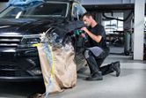 A person wearing safety equipment uses a polisher on a car fender in an auto shop.