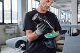 A person wearing safety equipment applies polish to a cordless polisher in an auto workshop.