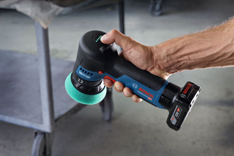A person holds a cordless polisher with a green foam pad near a metal cart.