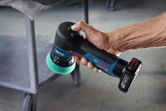 A person holds a cordless polisher with a green foam pad near a metal cart.