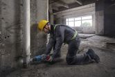 A person wearing safety equipment uses a reciprocating saw to cut a pipe near a concrete wall.