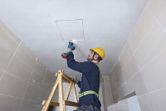 A person wearing safety equipment uses a reciprocating saw to cut a ceiling panel.