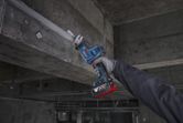 A person wearing safety equipment uses a reciprocating saw to cut metal overhead.