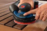 A person operates a cordless random orbit sander on a wooden surface.