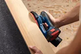 Person sanding a wooden plank using a cordless random orbit sander.