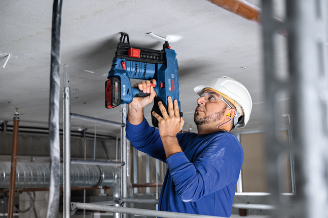 A person wearing safety equipment uses a cordless tool to fasten material to the ceiling.
