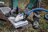 A person wearing safety equipment uses a large angle grinder with dust extraction to cut concrete.