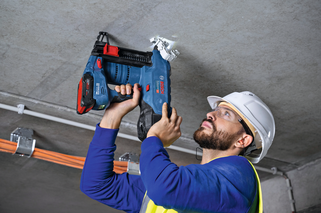 A person wearing safety equipment fastens a ceiling conduit with a cordless nail gun.