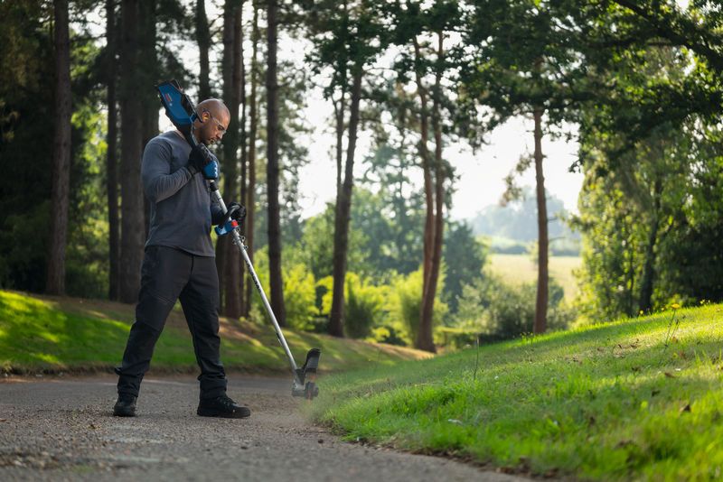 A person wearing safety equipment trims grass along a path with a cordless grass trimmer.