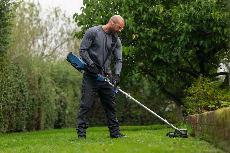 A person wearing safety equipment trims grass along a garden edge with a cordless tool.