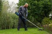 A person wearing safety equipment trims grass along a garden edge with a cordless tool.