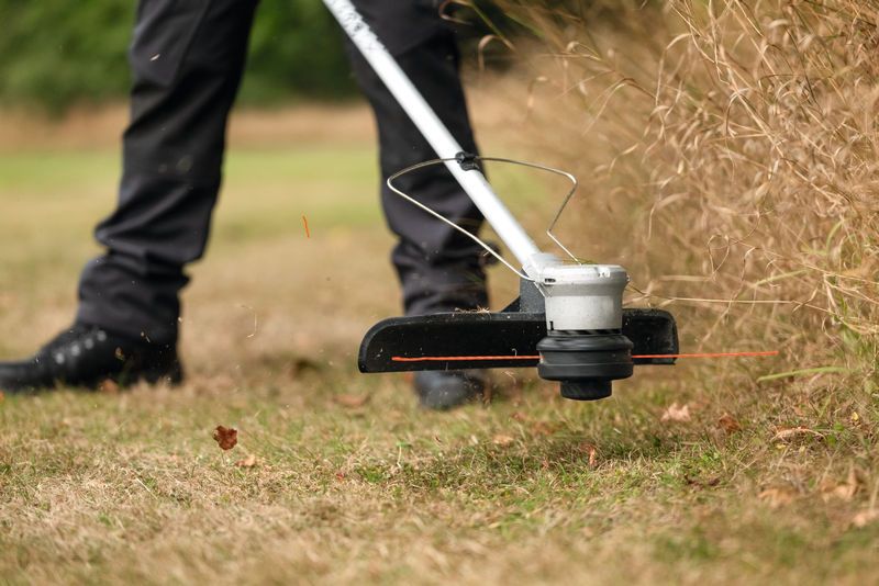 A person trims tall grass along a lawn edge with a cordless grass trimmer.