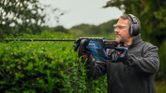 A person wearing safety equipment trims hedges with an electric hedge trimmer.