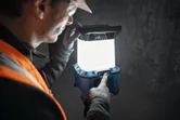 A person wearing safety equipment adjusts a cordless jobsite light in a dim workspace.