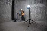 A person wearing safety equipment drills a concrete wall with a vacuum and standing jobsite light.
