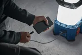 Person charging a smartphone using a cordless jobsite light on a concrete floor.