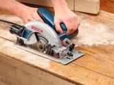 A person operates a hand-held circular saw, cutting wood on a workbench.