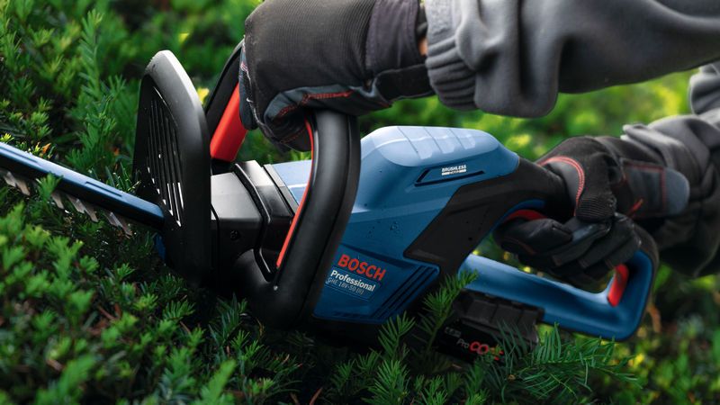 A person wearing safety equipment trims bushes with a blue cordless hedgecutter.