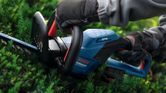 A person wearing safety equipment trims bushes with a blue cordless hedgecutter.