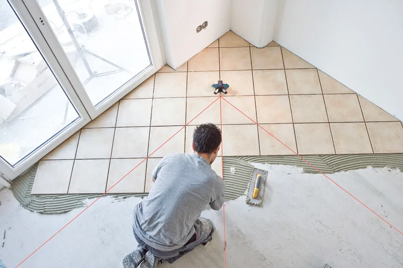 Person kneeling and aligning tile using a laser leveling tool.