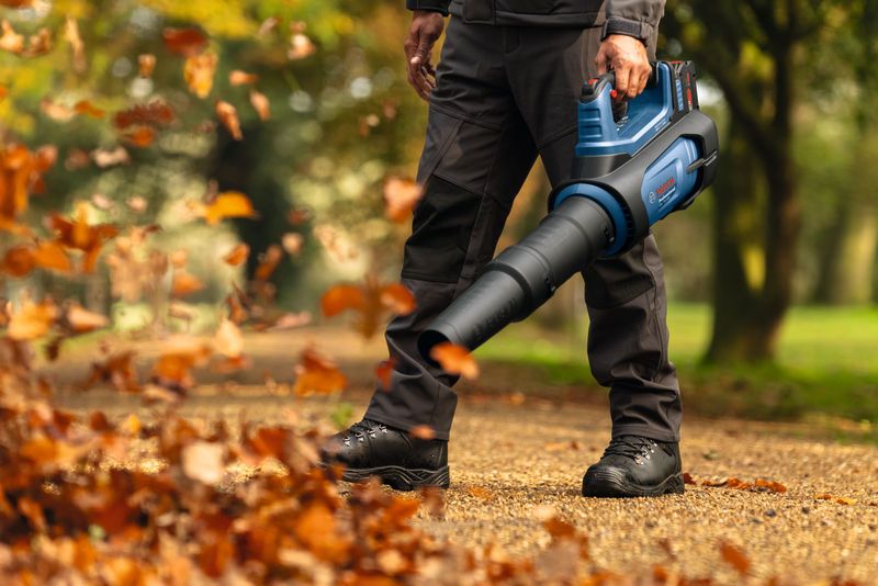 A person wearing safety equipment uses a cordless leaf blower to clear fallen leaves on a path.