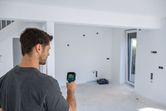 A person uses a thermo camera to scan a wall in an unfinished room.