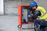 A person wearing safety equipment uses a digital multimeter to test an electrical panel.