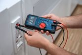 A person tests a wall electrical outlet using a digital multimeter.