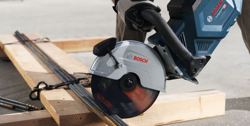 A person wearing safety equipment cuts metal rebar with a cordless grinder.