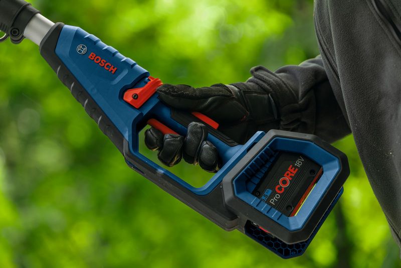 Person wearing safety equipment holds a cordless pole saw outdoors.