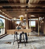 A person wearing safety equipment uses a planer to smooth a wooden beam in a building frame.