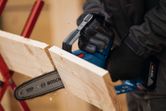 A person wearing safety equipment uses a cordless pruner saw to cut a wooden board.