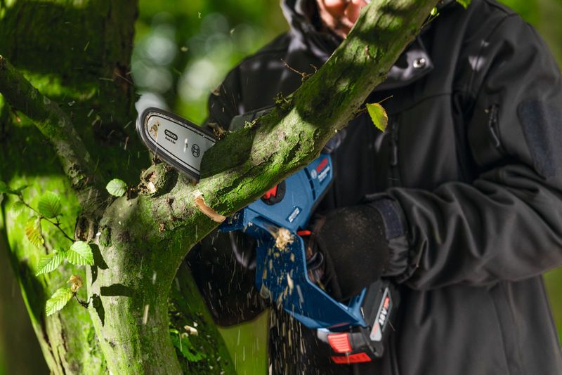 A person wearing safety equipment trims a tree branch with a cordless pruner saw.