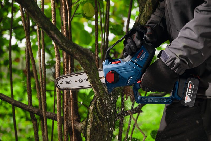 A person wearing safety equipment uses a cordless pruner saw to cut a tree branch.