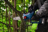 A person wearing safety equipment uses a cordless pruner saw to cut a tree branch.