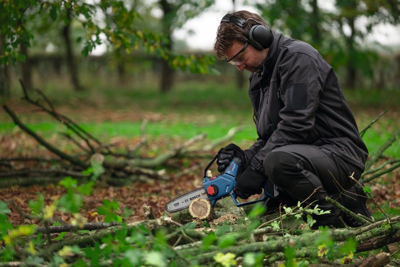 A person wearing safety equipment trims branches on the ground with a cordless pruner saw.