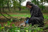 A person wearing safety equipment trims branches on the ground with a cordless pruner saw.