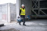 A person wearing safety equipment carries a metal cut-off saw in an unfinished building.