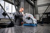 A person wearing safety equipment operates a metal cut-off saw in a workshop.