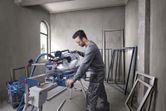 A person wearing safety equipment operates a mitre saw to cut metal frames indoors.