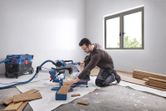 A person wearing safety equipment cuts wood planks with a mitre saw in a bright room.