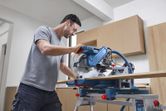 A person wearing safety equipment cuts wood with a mitre saw in a workshop.